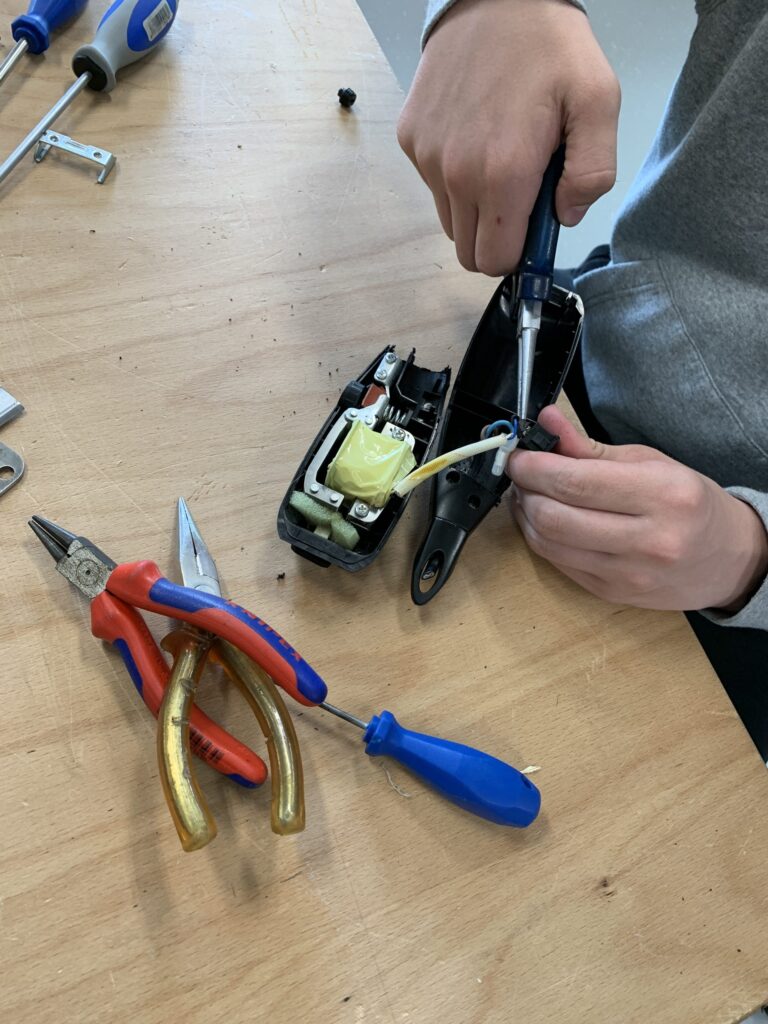 Close-up van een werktafel waarop een klein elektrisch apparaat is opengemaakt en wordt bewerkt met een schroevendraaier, naast tangen en ander gereedschap.